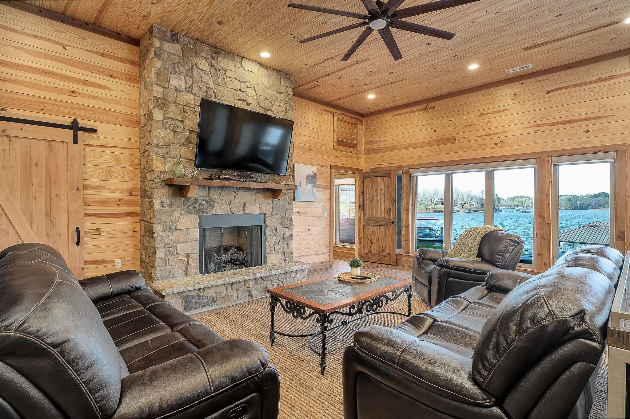 A cozy living room with a stone fireplace, leather sofas, and a large TV mounted on the wall.