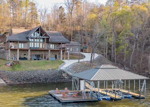 Une grande maison en pierre avec un quai qui s’avance sur un lac, entourée d’arbres.