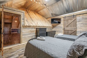Un intérieur en bois avec un plafond en pente, un lit recouvert d’une couverture grise, une commode et un téléviseur fixé au mur.