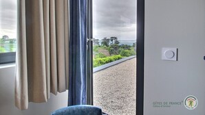 A room with a large sliding glass door leading to a balcony with a gravel surface and a view of trees and the sea. - La Roche Mignonne - Trebeurden (TREBEURDEN)