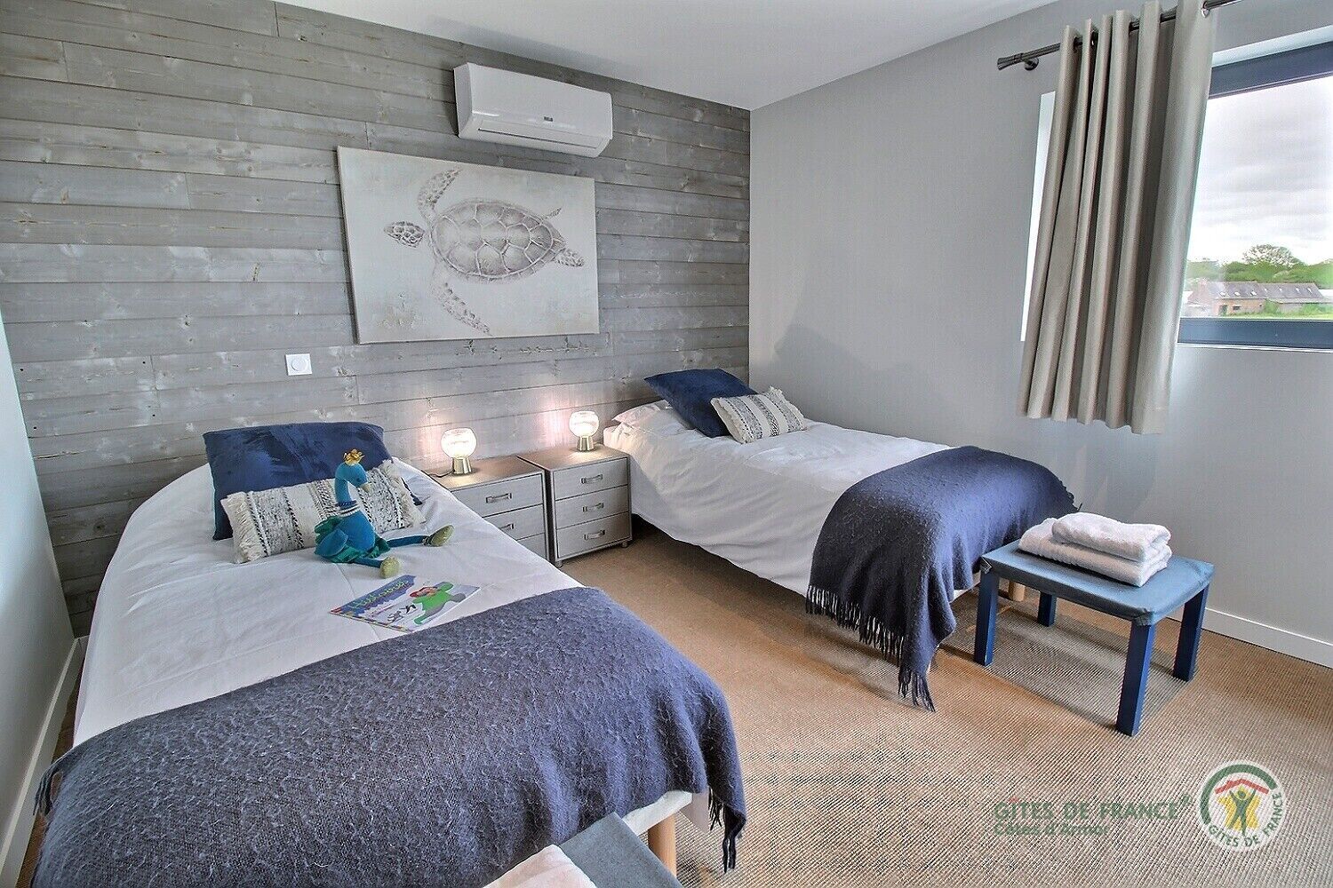 A bedroom with two beds, a wall-mounted air conditioner, a framed artwork, bedside lamps, and a view of the outdoors through a window with curtains.