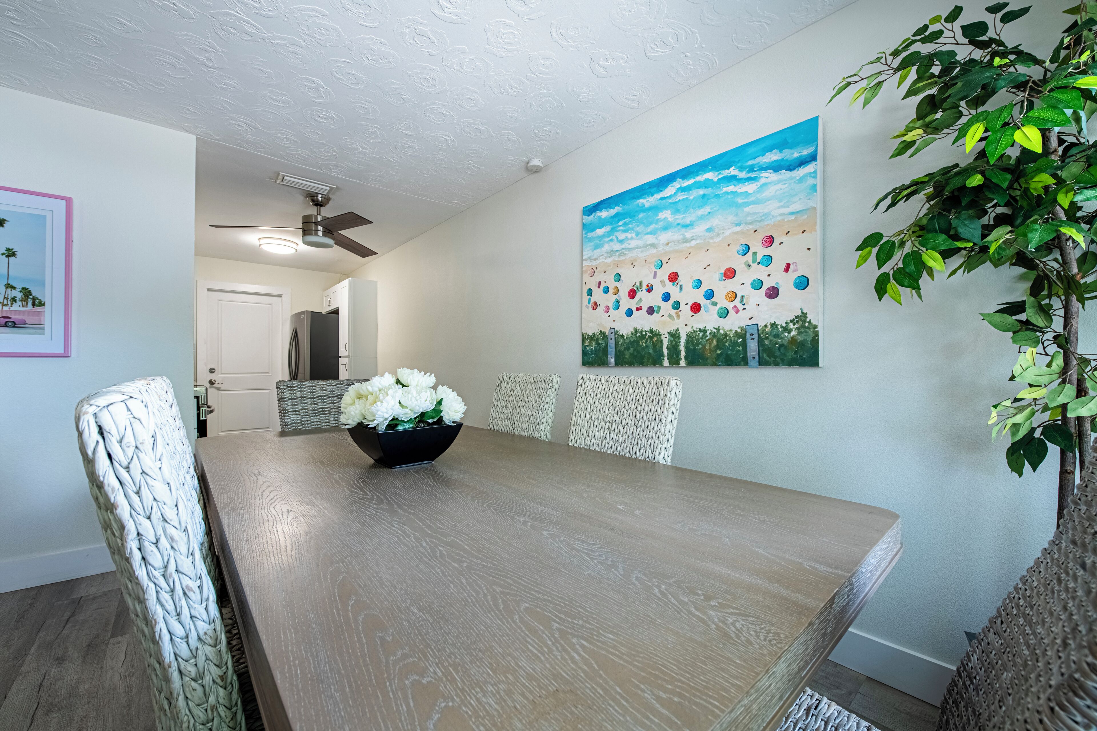 Un coin repas avec une table en bois, des chaises blanches, un ventilateur de plafond et un tableau accroché au mur.