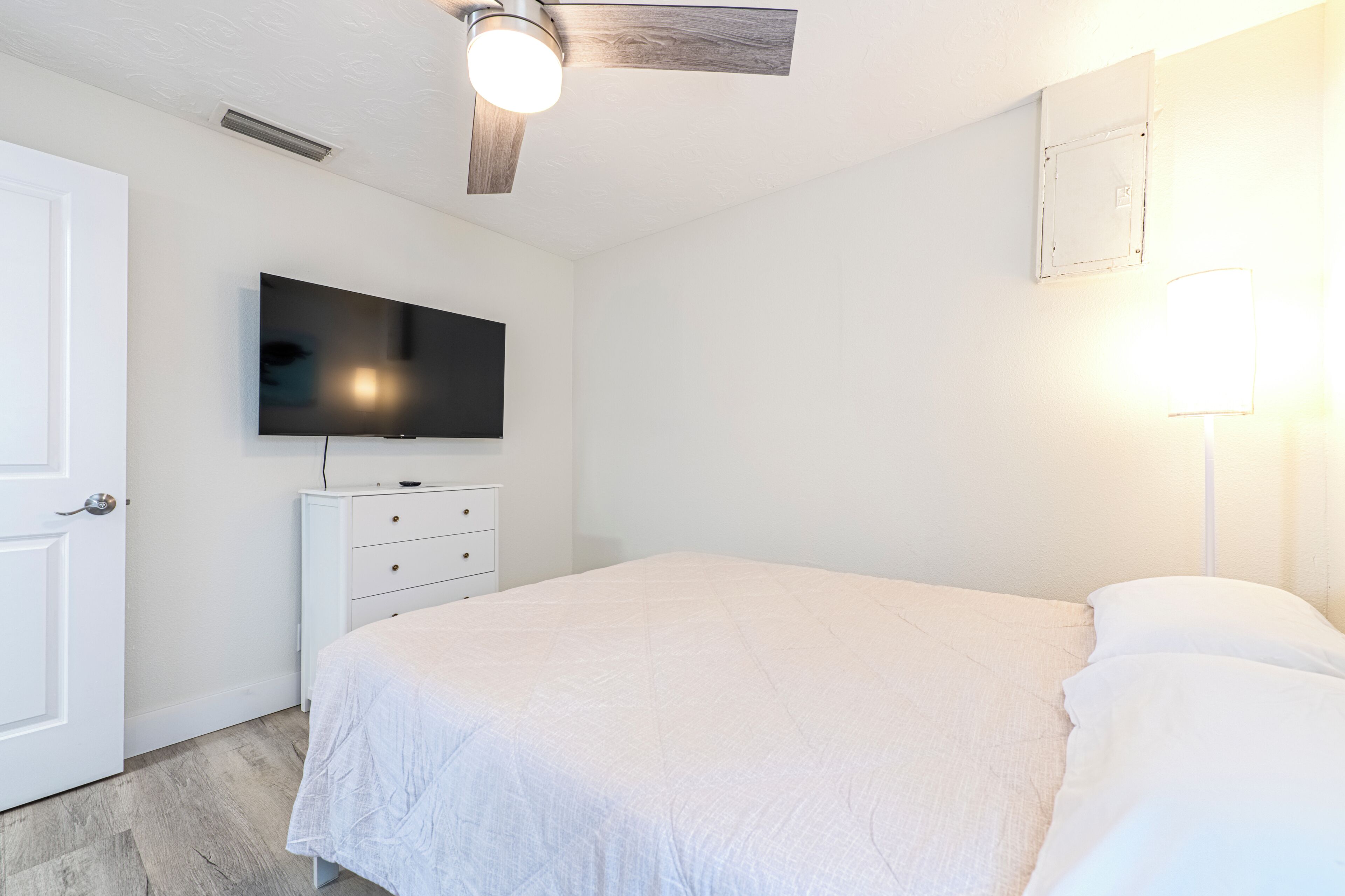Un dormitorio con cama, televisión montada en la pared, ventilador de techo y una puerta blanca.