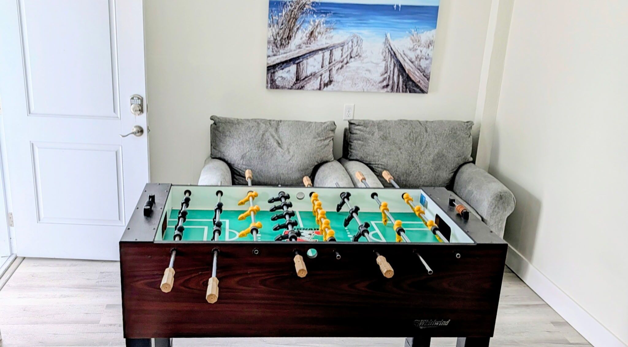 A foosball table in a room with a white door, grey sofa, and a painting on the wall.