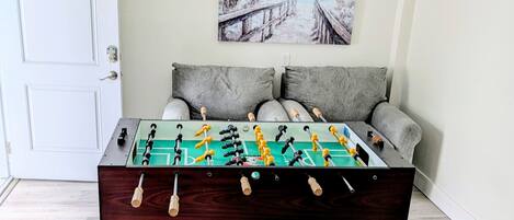 A foosball table in a room with a white door, grey sofa, and a painting on the wall.