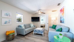 A living room with two grey sofas, a patterned ottoman, a green side table with a plant, and a ceiling fan. - Family-friendly 2-bed, 2-bath ground-floor unit in Indian Rocks Beach. Sleeps eight with full kitchen, WiFi, smart TV, beach gear, and direct pool access. A comfortable, convenient base for sand days, sunsets, and easy coastal living (Indian Rocks Beach)