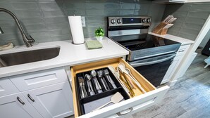 Una cocina con armarios blancos, un lavamanos de acero inoxidable, un horno empotrado y un cajón con utensilios.