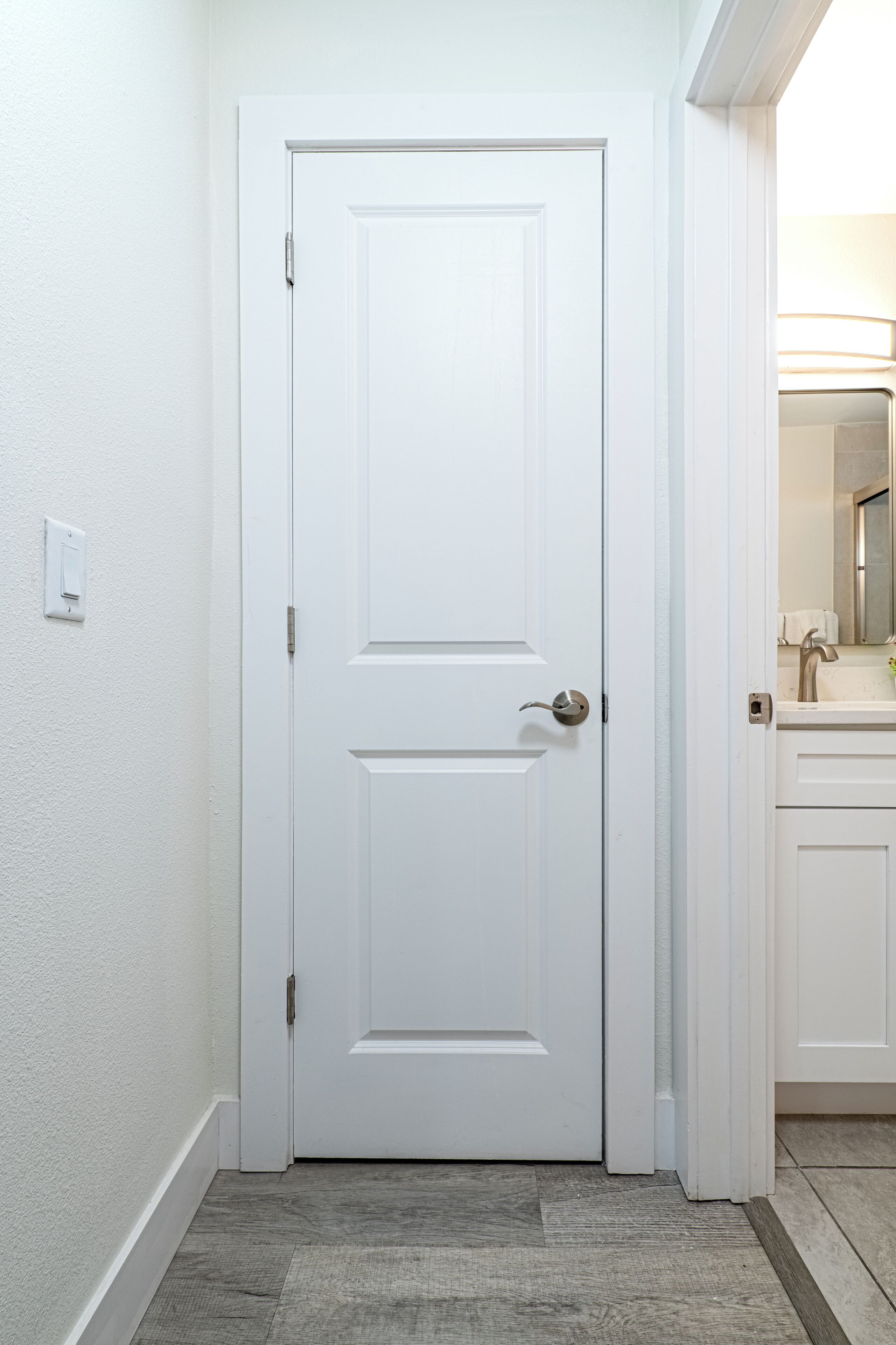 Une porte blanche avec une poignée argentée, un interrupteur sur le mur et une salle de bain avec un lavabo et un miroir visibles à travers l’ouverture de la porte.
