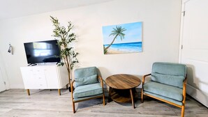 A living room with a TV on a white cabinet, two striped armchairs, a round wooden table, a plant, and a beach-themed painting on the wall. - MySunsetView 6 Unit 4 Studio w kitchenette (Indian Rocks Beach)