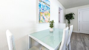 Ein Essbereich mit einem Glastisch, weißen Stühlen, einer Topfpflanze und einem farbenfrohen Bild an der Wand.