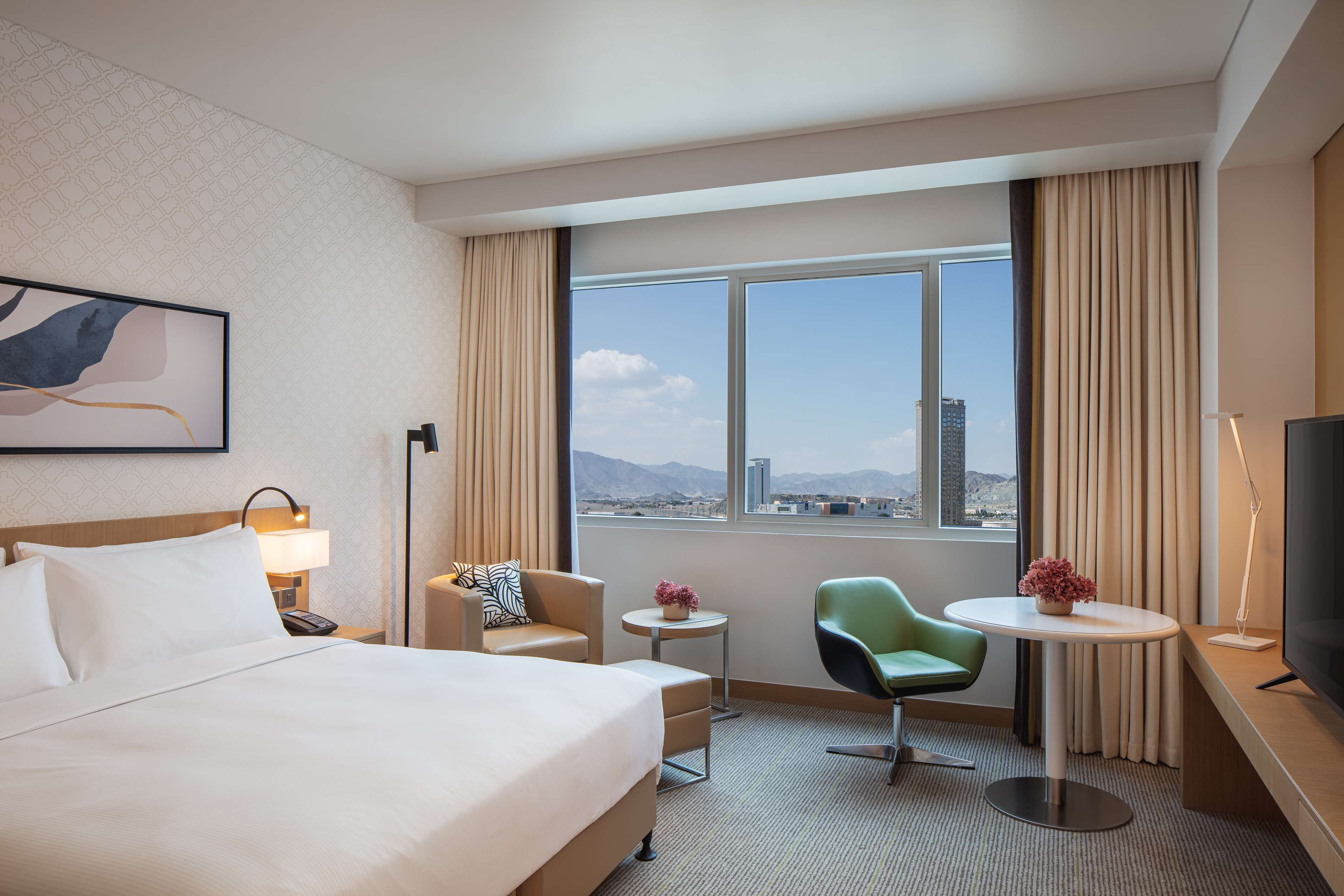 A hotel room with a large bed, a desk with a lamp, a chair, a small table with a flower, and a TV.