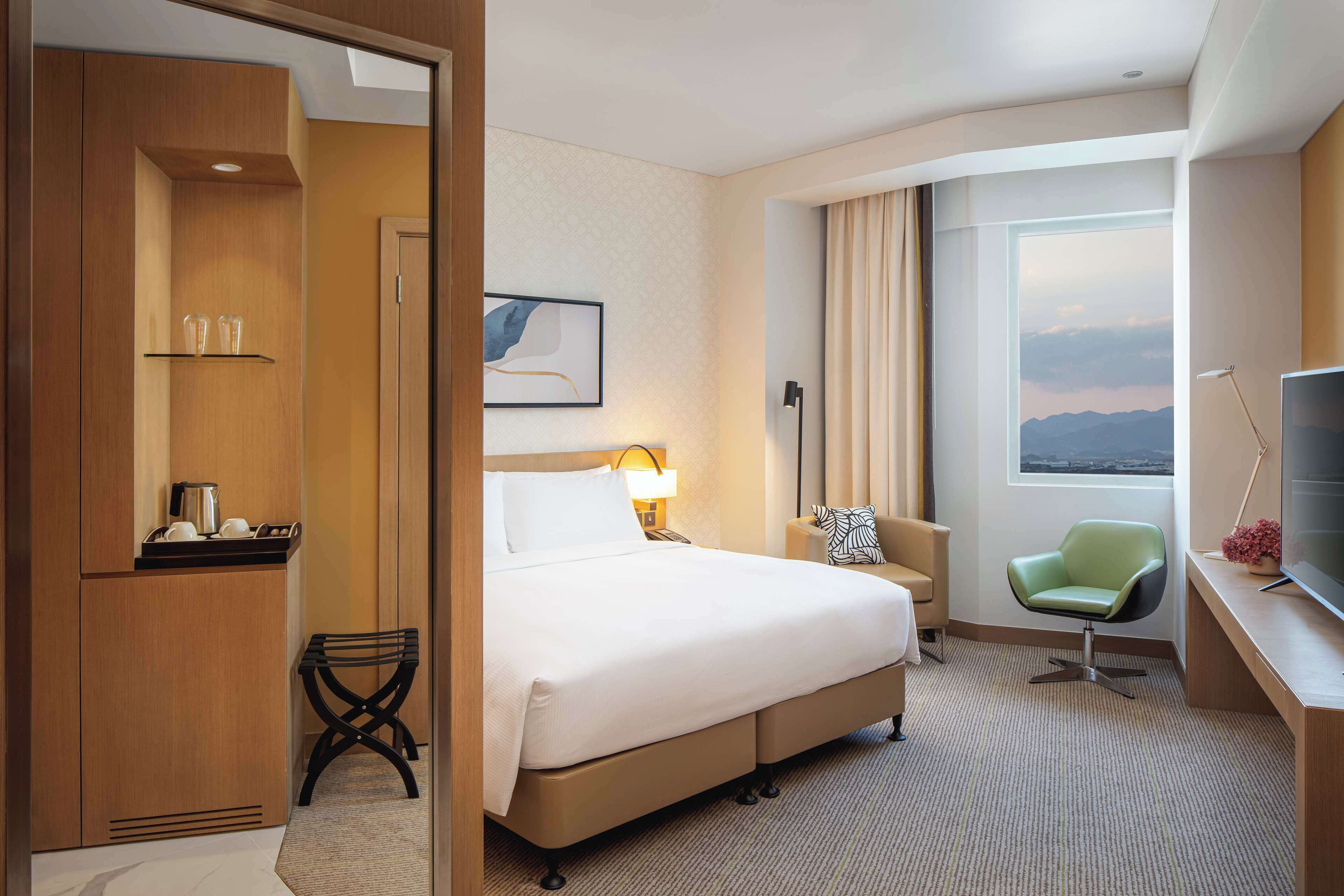 A hotel room with a bed, a desk with a television, a chair, a desk lamp, a window with a view, and a small shelf with glasses.
