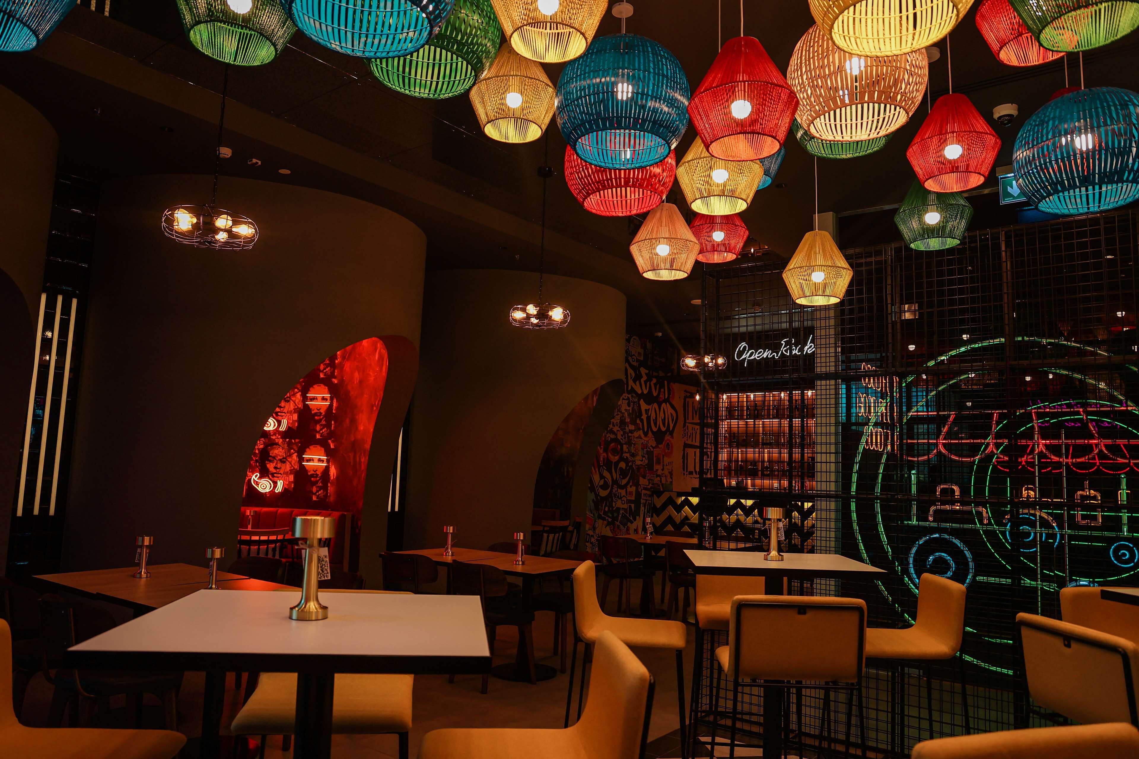 Ein Restaurantinneres mit mehreren Tischen und Stühlen, bunten hängenden Lampen und einem Neon-Schild an der Wand.