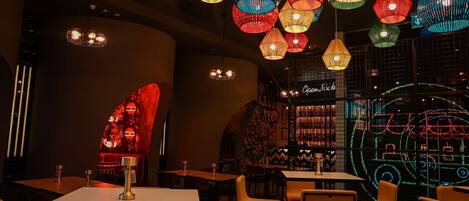 Ein Restaurantinneres mit mehreren Tischen und Stühlen, bunten hängenden Lampen und einem Neon-Schild an der Wand.