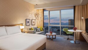Een hotelkamer met een groot bed, een sofa, een klein tafeltje en uitzicht op de stad door grote ramen.