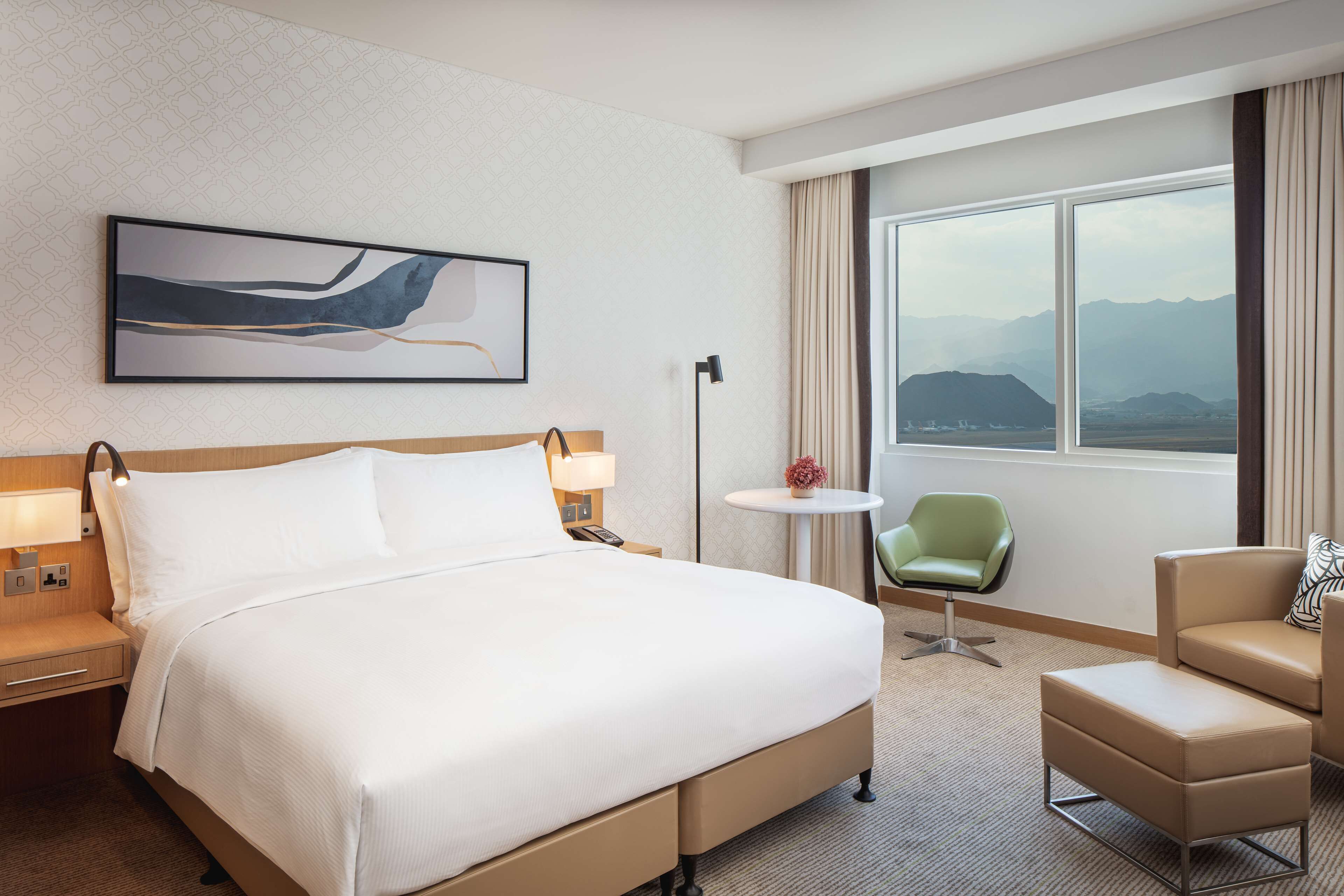 A hotel room with a large bed, a desk with a lamp, a small round table, a green chair, a beige armchair, and a view of mountains through the window.