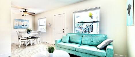 A living room with a teal sofa, a round coffee table, a dining area with a white table and chairs, and a ceiling fan.