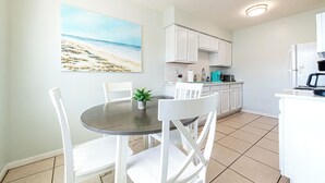 A dining area with a round table, white chairs, and a painting of a beach scene. - Cozy 2-bed, 1-bath second-floor unit minutes from Indian Rocks Beach. Sleeps 7 with full kitchen, WiFi, smart TV, beach gear, parking, and direct pool access. A bright, comfortable stay for families and groups (Indian Rocks Beach)