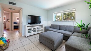 A modern living room with a grey sectional sofa, a flat-screen TV on a white cabinet, a large window with a view of palm trees, and a tiled floor. - Cozy 2-bed, 1-bath second-floor unit minutes from Indian Rocks Beach. Sleeps 7 with full kitchen, WiFi, smart TV, beach gear, parking, and direct pool access. A bright, comfortable stay for families and groups (Indian Rocks Beach)