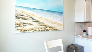 A painting of a beach scene on a wall, a white chair, and a kitchen area with cabinets and a trash bin. - Cozy 2-bed, 1-bath second-floor unit minutes from Indian Rocks Beach. Sleeps 7 with full kitchen, WiFi, smart TV, beach gear, parking, and direct pool access. A bright, comfortable stay for families and groups (Indian Rocks Beach)