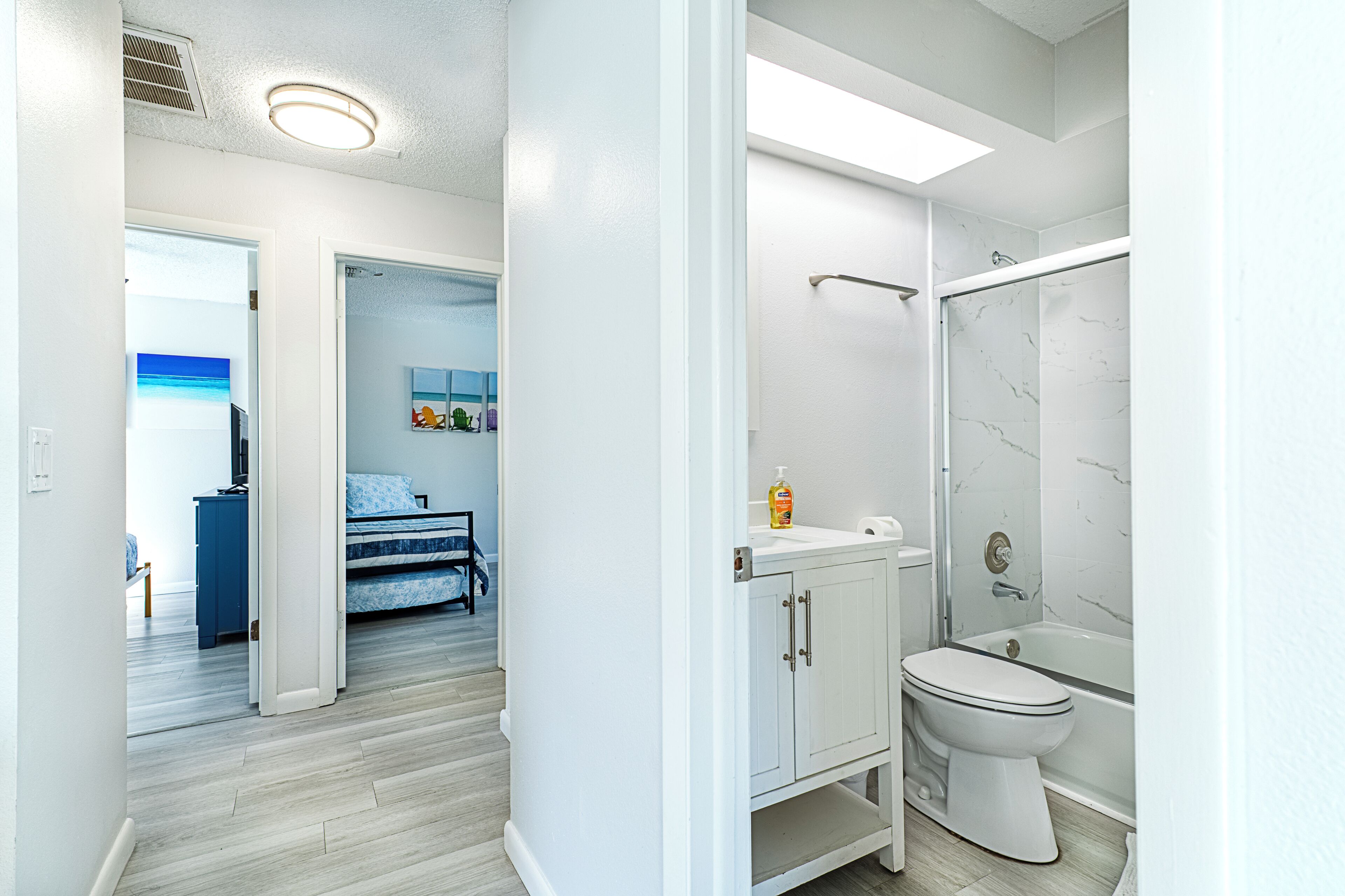 A modern bathroom with a shower, toilet, and sink. There is a bed in the adjacent room.