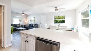 A modern kitchen with white cabinetry, a dishwasher, and a large island. There is a dining area with white chairs and a table, and a living room with a sofa and a coffee table.