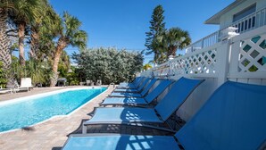 A poolside area with lounge chairs aligned along the pool, surrounded by palm trees and a white fence.