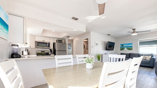 A modern kitchen with white cabinetry, a dining area with a wooden table and white chairs, and a living room with a grey sofa and a TV mounted on the wall.