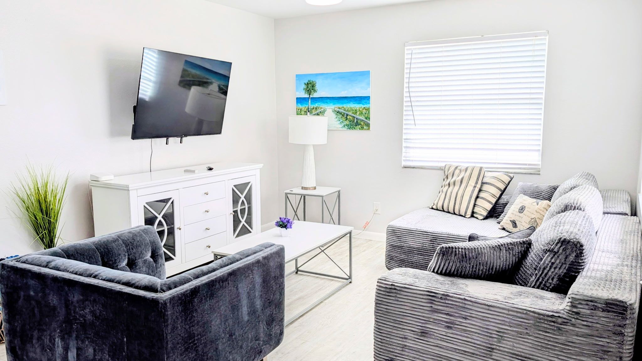 Un salon avec un téléviseur à écran plat, un tableau représentant une plage, une lampe et un canapé agrémenté de coussins à rayures.