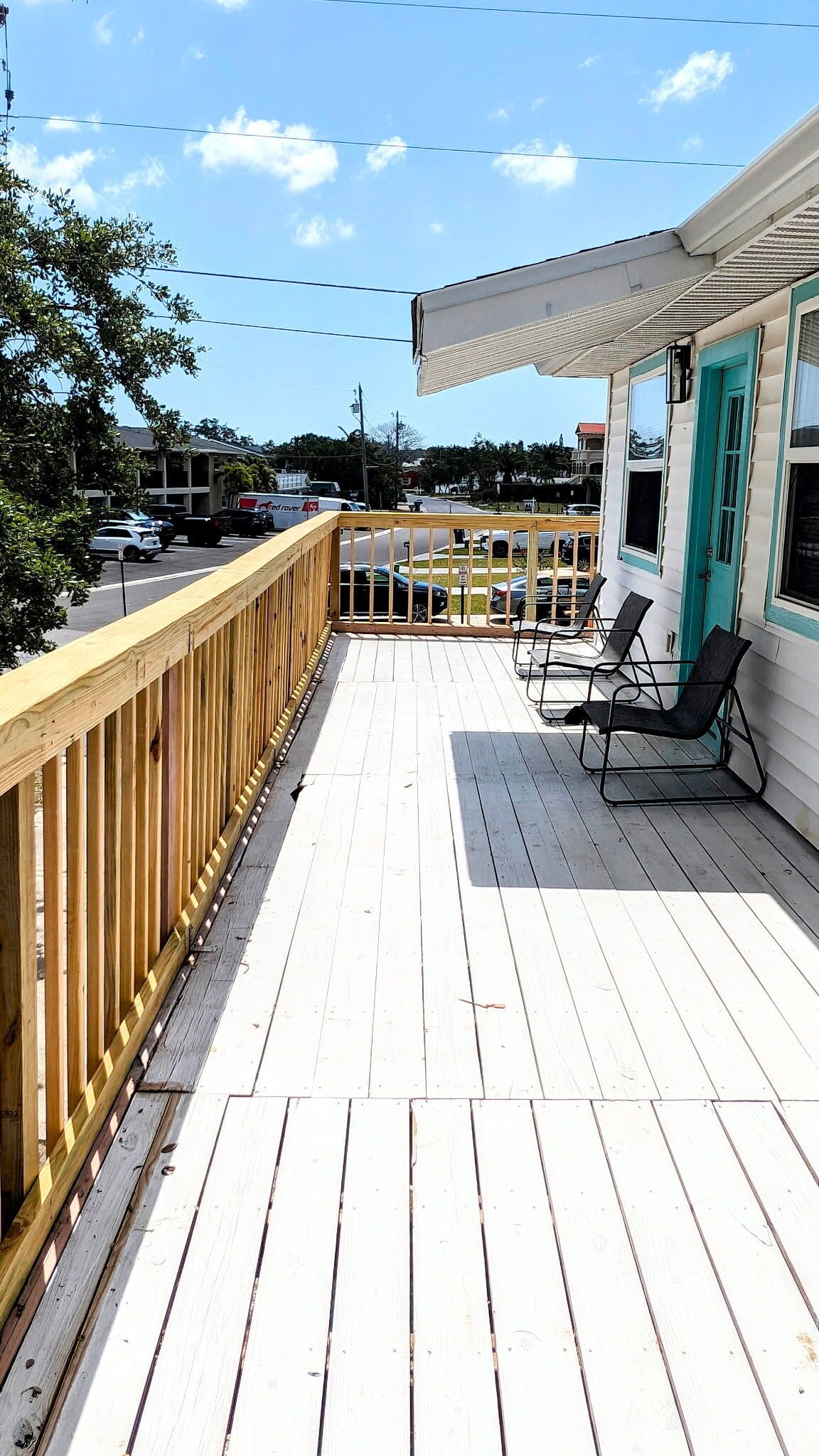 A wooden deck with a railing, a covered area with two chairs, and a building with a teal door.