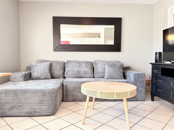 A modern living room with a sectional sofa, a round wicker table, a dark wooden cabinet, and a large mirror on the wall.