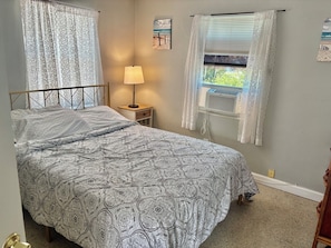 A bedroom with a bed, a nightstand with a lamp, an air conditioning unit, and a window with a view.