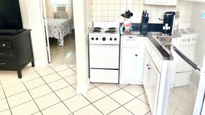 A compact kitchen with white appliances, a black TV stand, and a bed visible through an open door.