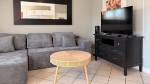 A living room with a grey sofa, a round wooden table, a black TV stand, and a large mirror.