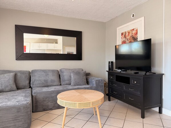 A living room with a grey sofa, a round wooden table, a black TV stand, and a large mirror.