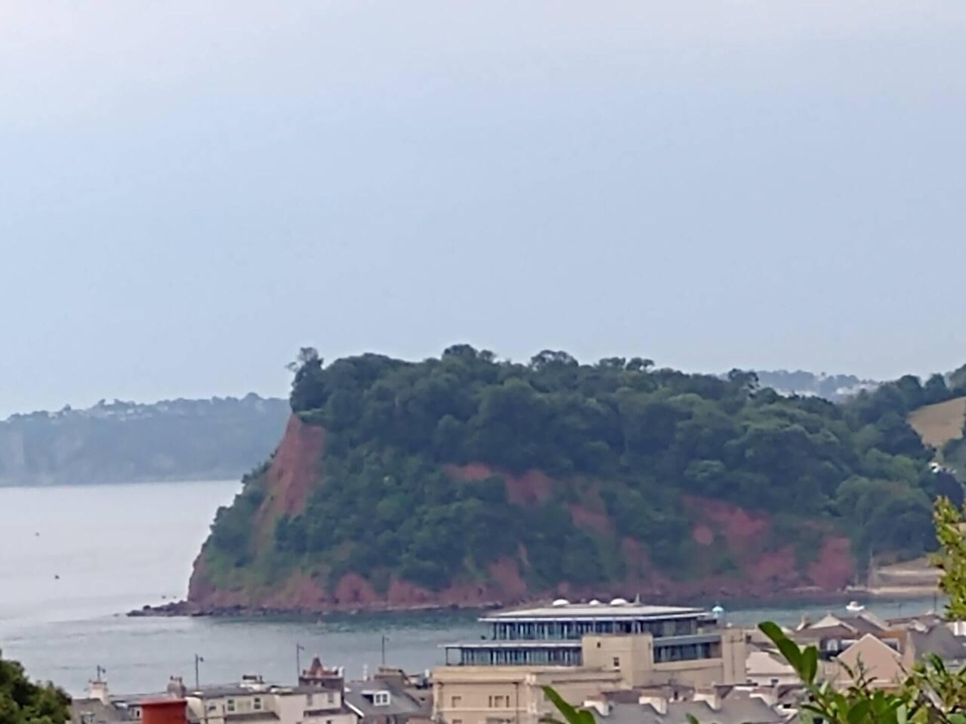 A coastal town with buildings near the water, a prominent red cliff, and a large building with a flat roof.