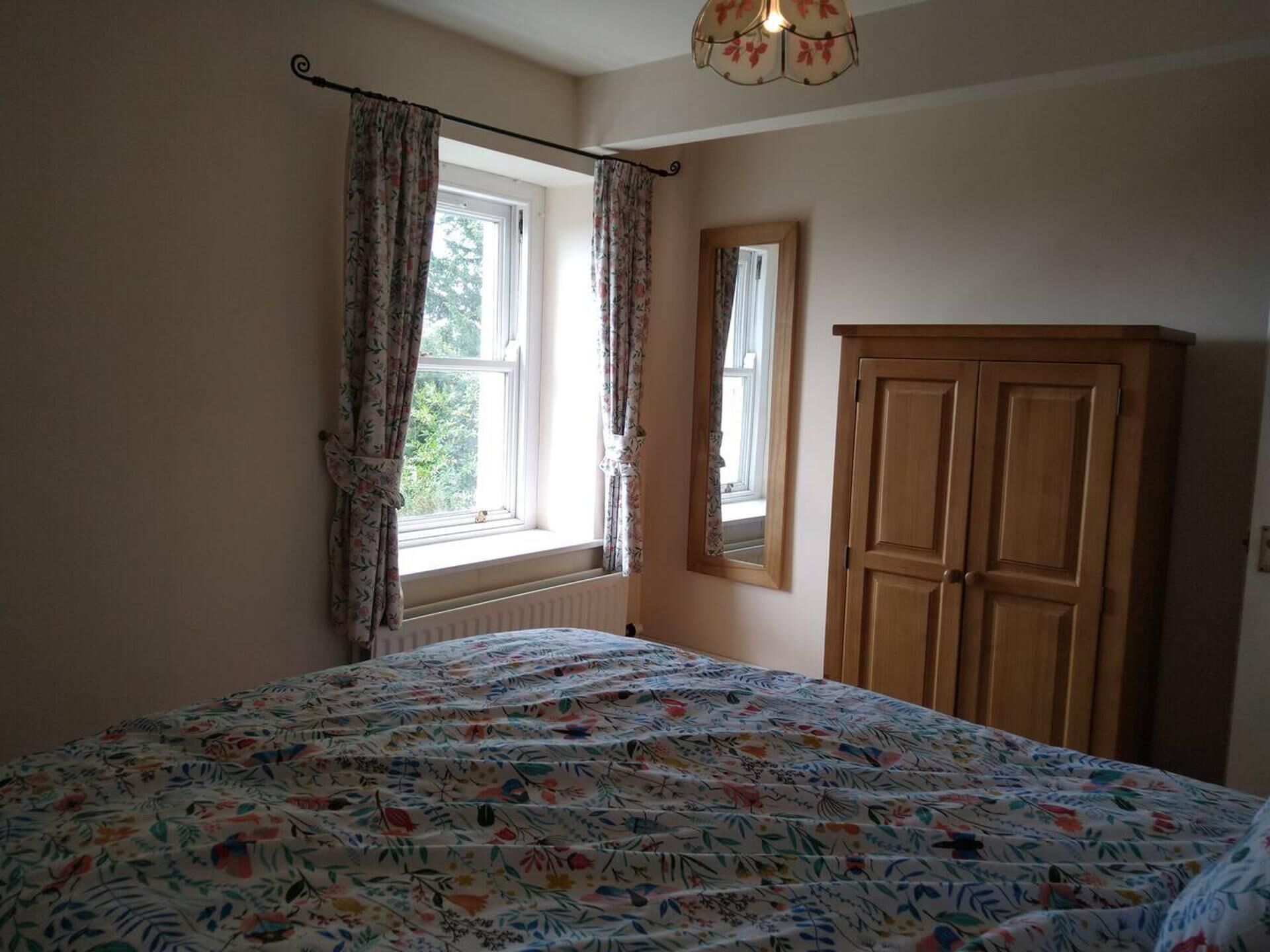 A bedroom with a floral-patterned bedspread, a wooden wardrobe, a window with curtains, and a mirror on the wall.