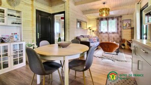 Een gezellige, goed verlichte kamer met een houten plafond, een ronde eettafel met stoelen, een bank en een keukenhoek met kasten en een raam.