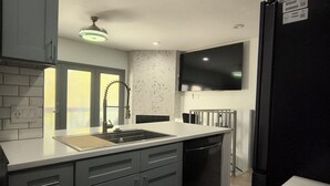 A modern kitchen with grey cabinets, a black refrigerator, a built-in dishwasher, and a large flat-screen TV mounted on the wall. - Urban Treehouse (Dallas)