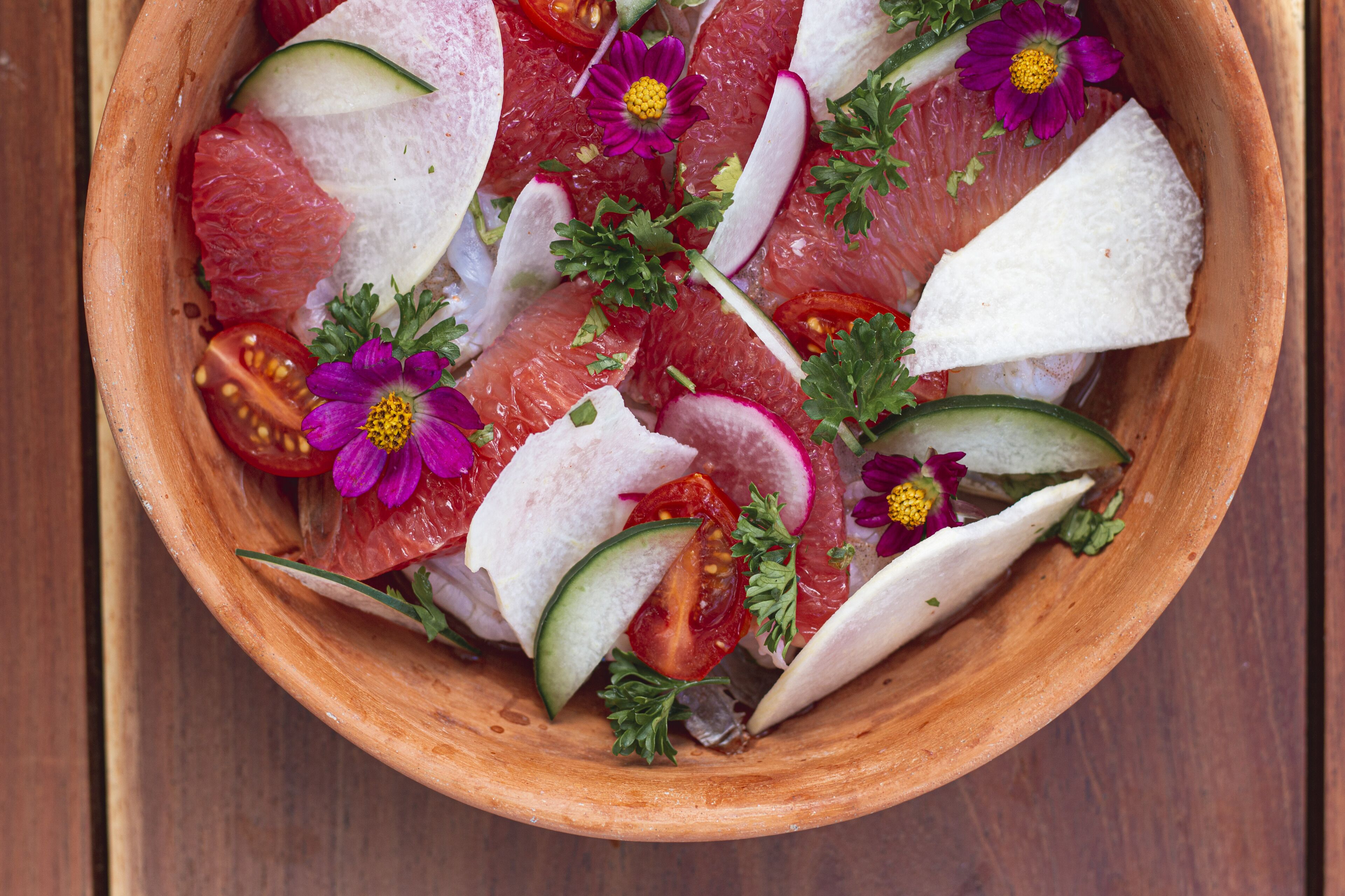 A wooden bowl filled with sliced fruits and vegetables, garnished with flowers.