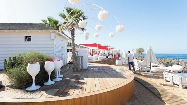 Eine Holzterrasse mit Weingläsern, einer Palme und einem Strand im Hintergrund.