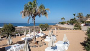 Ein Strand mit weißen Liegestühlen und Sonnenschirmen, Palmen und freiem Blick auf den Ozean.