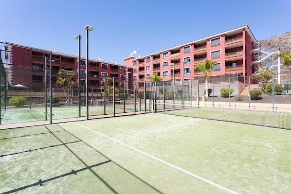 A tennis court with a net, surrounded by a fence, and adjacent to a multi-story building with balconies.