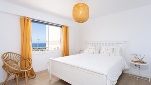 A bedroom with a large bed, a wicker chair, a bedside table, and a view of the sea through the window. - Home2Book Beachfront Design&Views Los Cristianos (Los Cristianos)