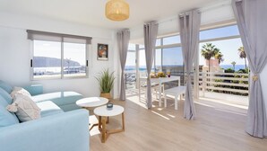 A modern living room with a sofa, two small tables, a hanging light fixture, and large windows offering a view of the sea and palm trees. - Home2Book Beachfront Design&Views Los Cristianos (Los Cristianos)