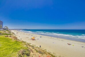 A beach with a pier extending into the ocean, sandy shores, and a grassy area with buildings in the background.