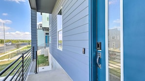 A modern building with a blue exterior, a covered outdoor area with a metal railing, and a clear view of a road and greenery in the background. - Luxurious & Spacious Austin Home ~ 8 Mi to Dtwn! (Austin)