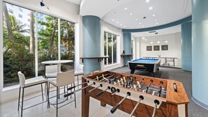 Un espace de loisirs intérieur moderne, doté d’un baby-foot, d’une table de billard et d’un coin salon avec de grandes fenêtres donnant sur un paysage verdoyant.