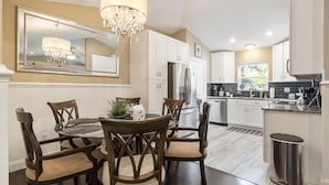 A modern kitchen with white cabinets, a stainless steel refrigerator, and a dining area with wooden chairs and a glass table. - Snow Birds Welcome. 10 min to Downtown & Beach (St. Augustine)