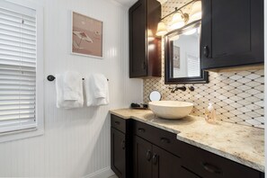 Une salle de bain avec un meuble-lavabo blanc, des armoires sombres, un grand miroir et un pare-feu en carreaux.